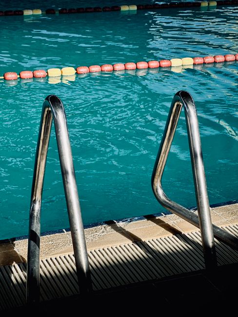 Close-up view of a swimming pool's metal ladder with two curved handrails leading into clear blue water. The pool features a floating lane rope with alternating pink and yellow buoys, indicating designated swimming areas. The poolside has a textured drainage grate along the edge, and sunlight reflects on the water's surface, casting subtle shadows. Inside a property, on the poolside, there are boxes, furniture wrapped in plastic or fabric, and packing materials arranged nearby, suggesting preparations for home relocation or furniture transport. The scene shows a typical residential or public swimming pool area in the context of moving or packing services provided by Removal Companies Tooting, with an emphasis on logistical considerations for efficient home removals or swimming pool equipment transport.