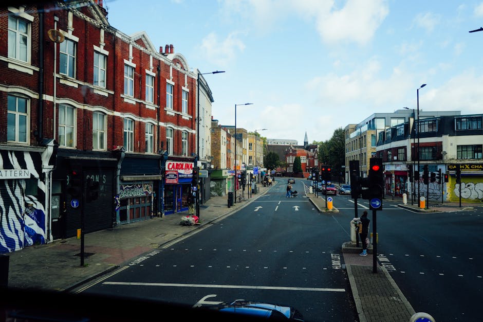 A street scene in Tooting with buildings on both sides, featuring a mix of residential and commercial properties. On the left, there are red brick and painted shopfronts, some with signage, and a few store entrances visible. The pavement is occupied by pedestrians, including a person standing near a traffic pole. The road has multiple lanes with white directional arrows, and traffic lights at the intersection are red. On the right, modern buildings with large windows and balconies are visible, along with a woman walking on the pavement near a street light. The sky is partly cloudy with blue skies and sunlight illuminating the scene. Vehicles are parked along the curbs, and some are visible on the road, indicating ongoing or upcoming vehicle movement. This urban environment is suitable for house removals, furniture transport, and home relocation processes, demonstrating typical street logistics encountered during moving services provided by Removal Companies Tooting.
