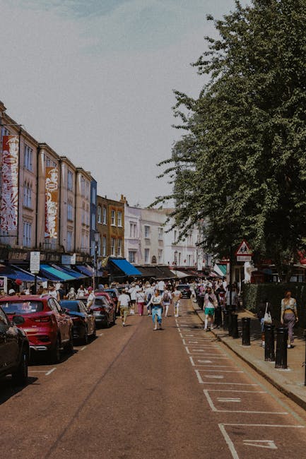 A street scene in Tooting with buildings on both sides, featuring a mix of residential and commercial properties. On the left, there are red brick and painted shopfronts, some with signage, and a few store entrances visible. The pavement is occupied by pedestrians, including a person standing near a traffic pole. The road has multiple lanes with white directional arrows, and traffic lights at the intersection are red. On the right, modern buildings with large windows and balconies are visible, along with a woman walking on the pavement near a street light. The sky is partly cloudy with blue skies and sunlight illuminating the scene. Vehicles are parked along the curbs, and some are visible on the road, indicating ongoing or upcoming vehicle movement. This urban environment is suitable for house removals, furniture transport, and home relocation processes, demonstrating typical street logistics encountered during moving services provided by Removal Companies Tooting.