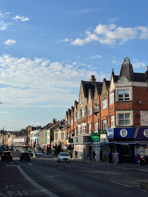 A residential street during daytime with rows of parked cars lining both sides of the narrow tarmac road, which is used for house removals and home relocation services. The street features typical ter